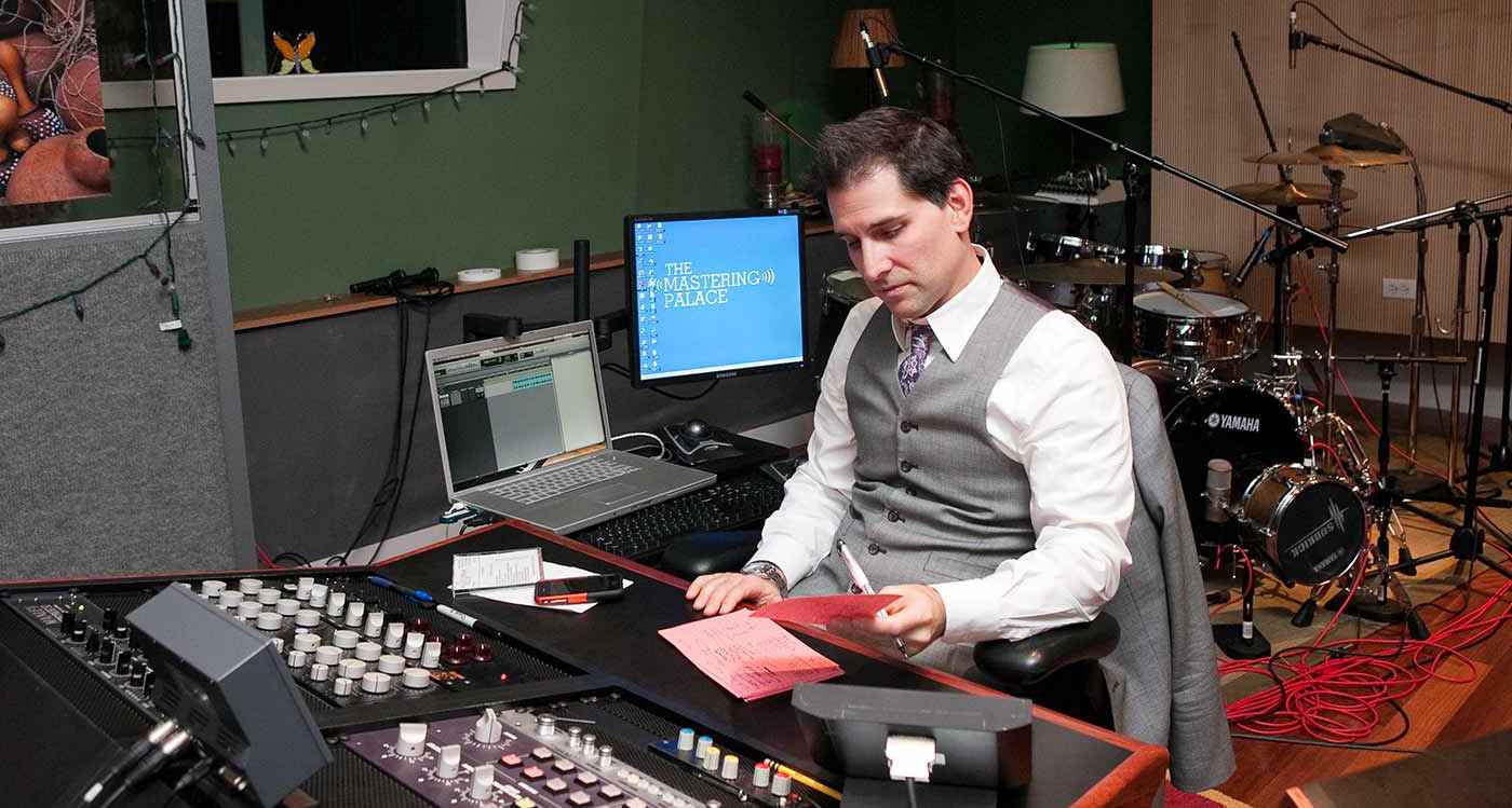 Dave Kutch sat in a suit in a recording studio, with a drum set behind him and a pen and paper with notes on that he is looking at