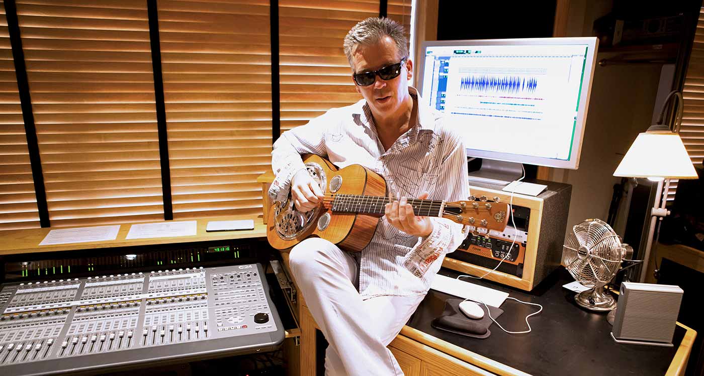 Simon Climie playing an acoustic guitar in a mixing studio in front of a computer screen with Pro Tools displayed